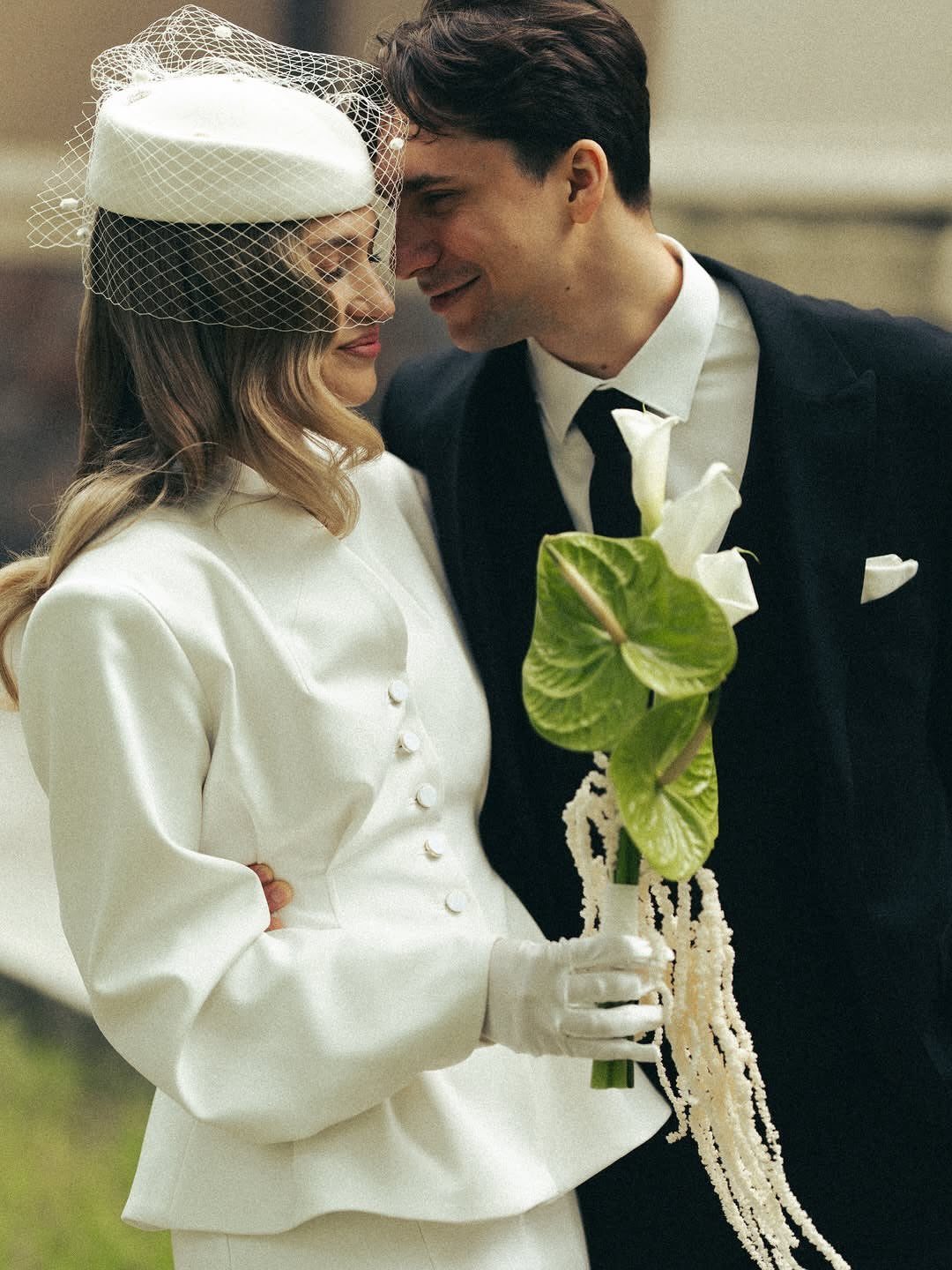 Couple in wedding attire, woman in white suit and pillbox hat holding lilies.