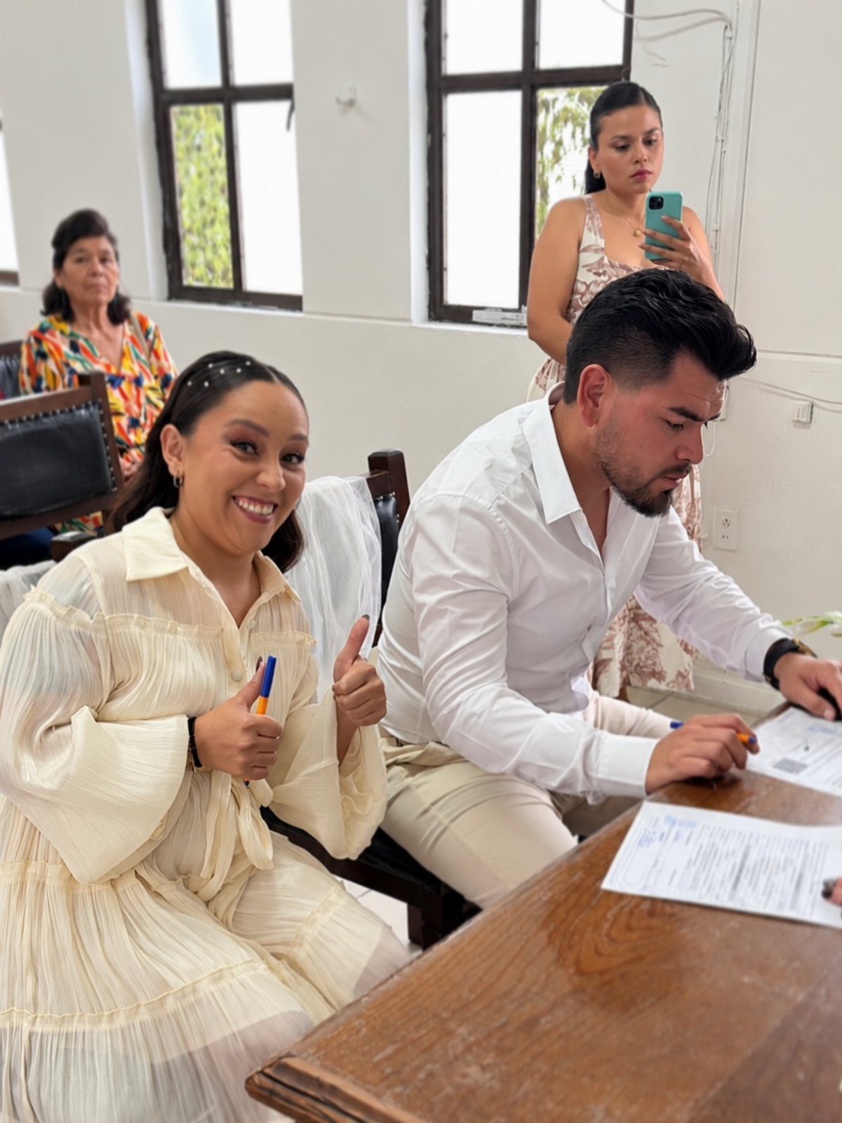 Couple at a wooden desk, woman smiling and man signing marriage documents.