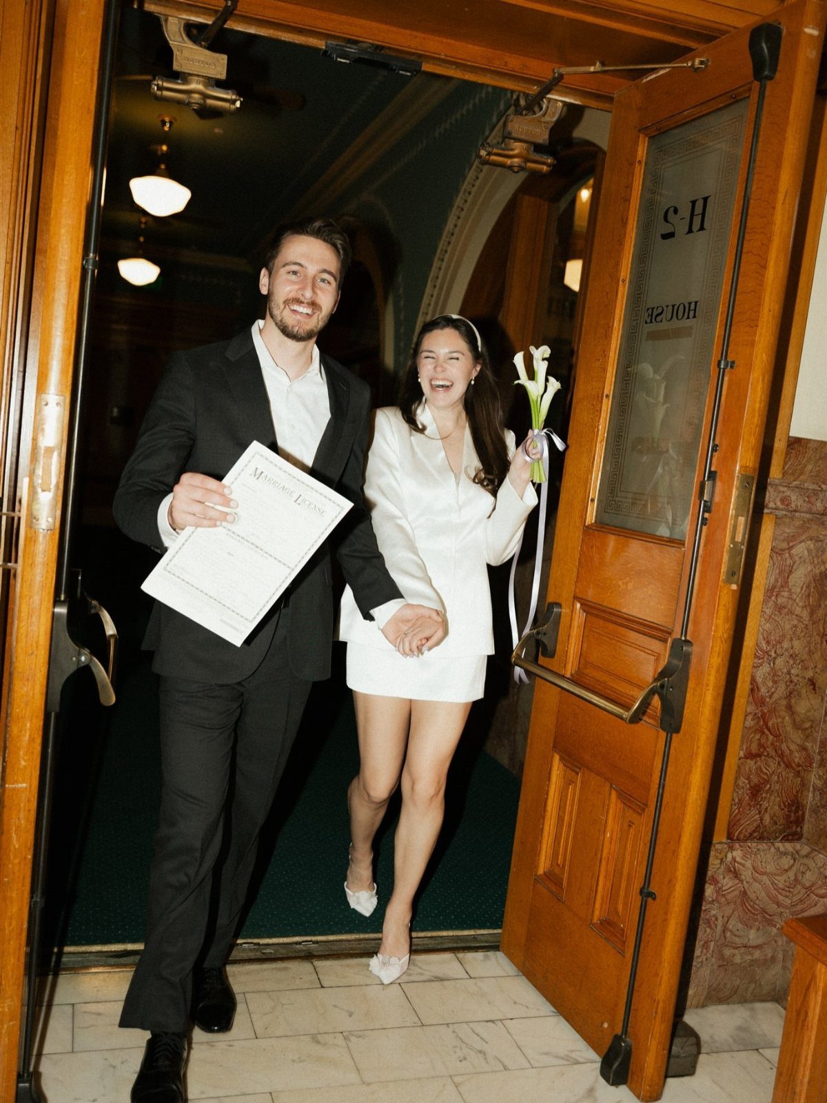 Newlywed couple smiling while exiting a building through large wooden doors.
