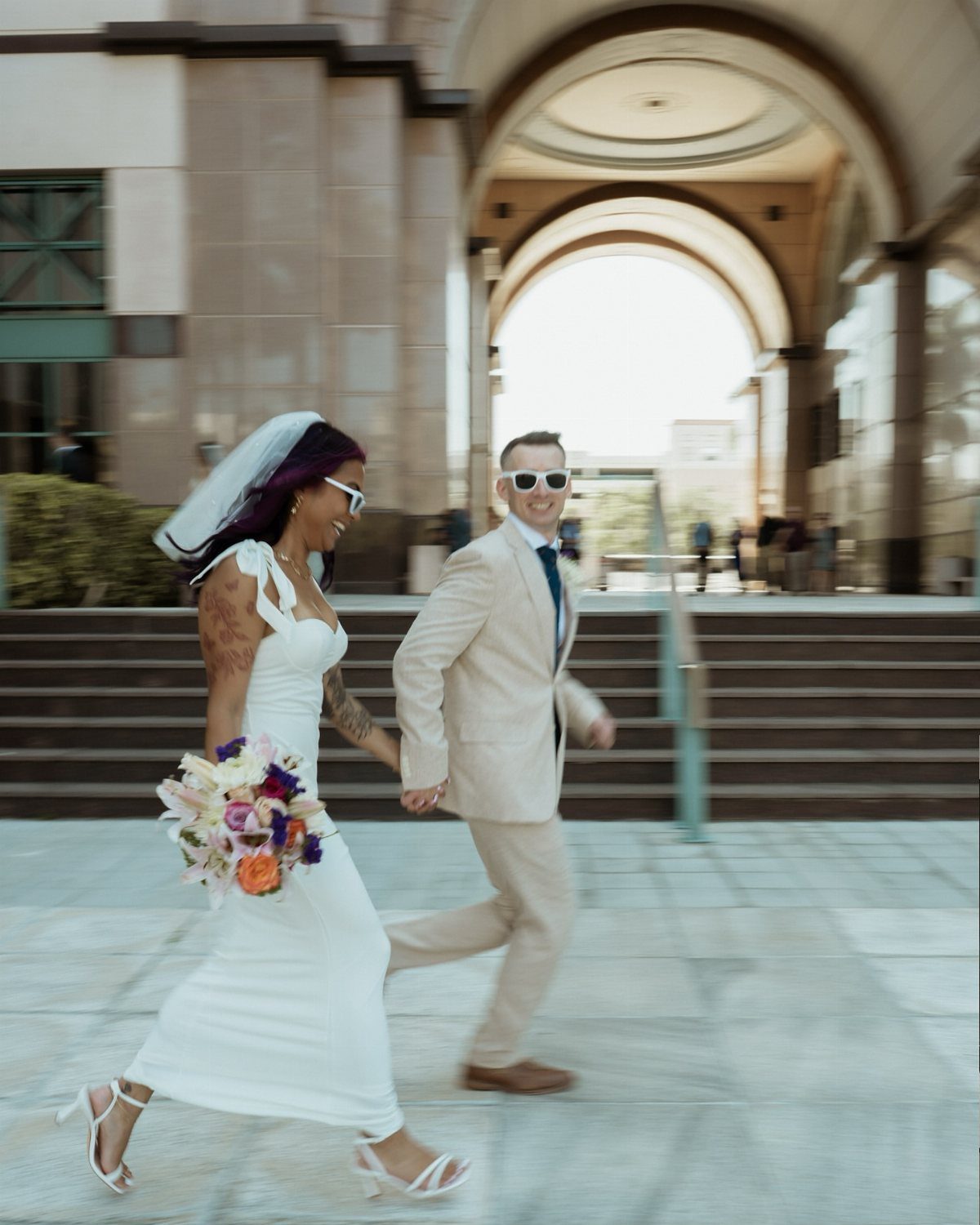 Bride and groom walking hand-in-hand in front of a courthouse with stone arches.