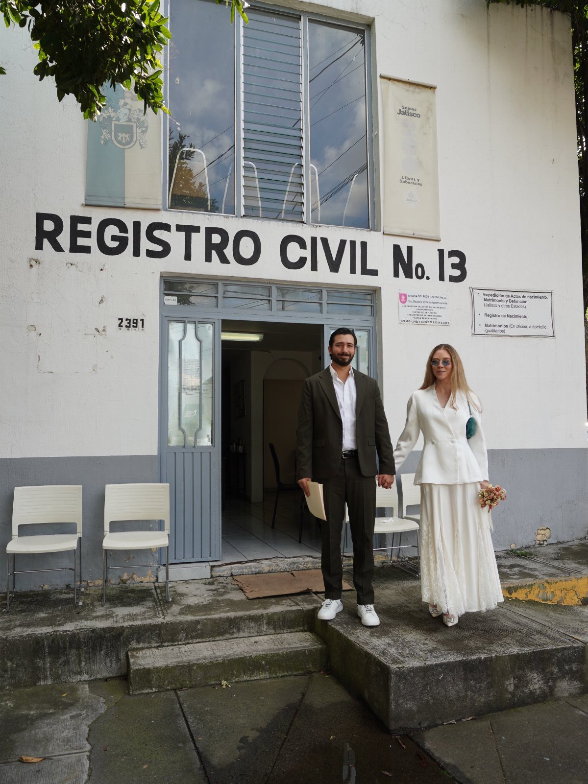 Couple in wedding attire posing in front of a civil registry building entrance.