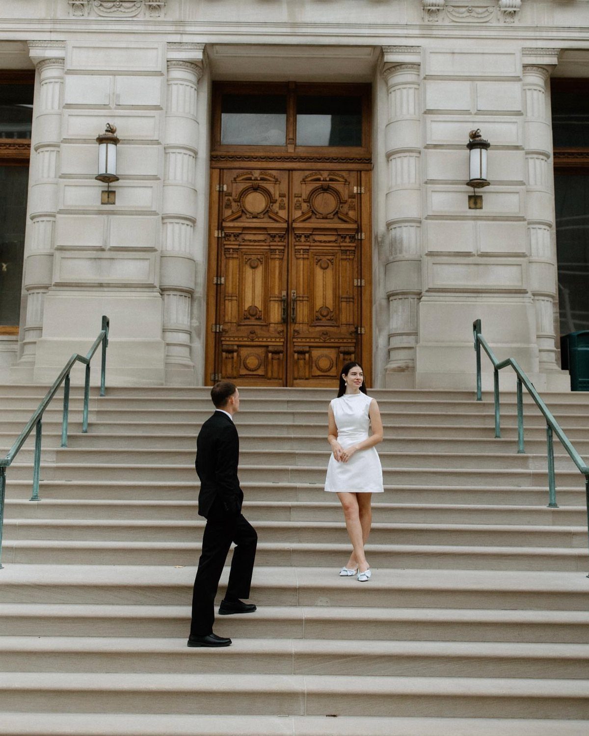 Couple in wedding attire standing on the stone steps of a courthouse building.