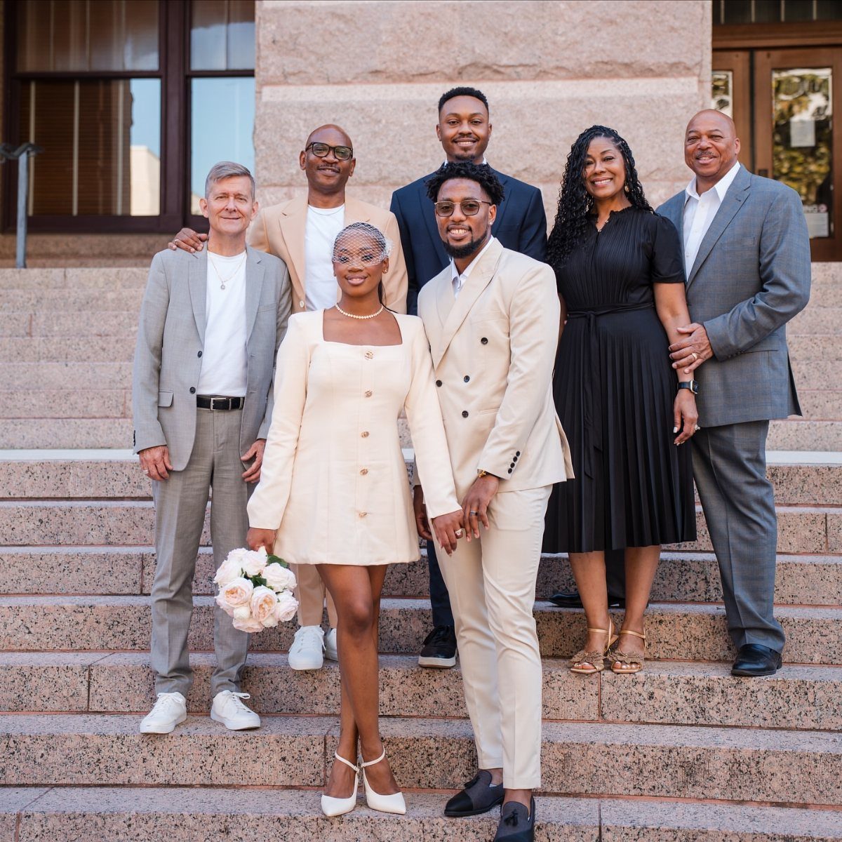 Newlywed couple in cream outfits posing with family on stone courthouse steps.