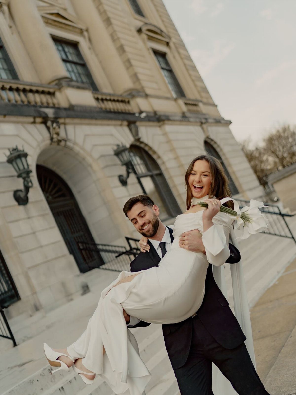 Groom carrying bride in white dress in front of a classical stone building