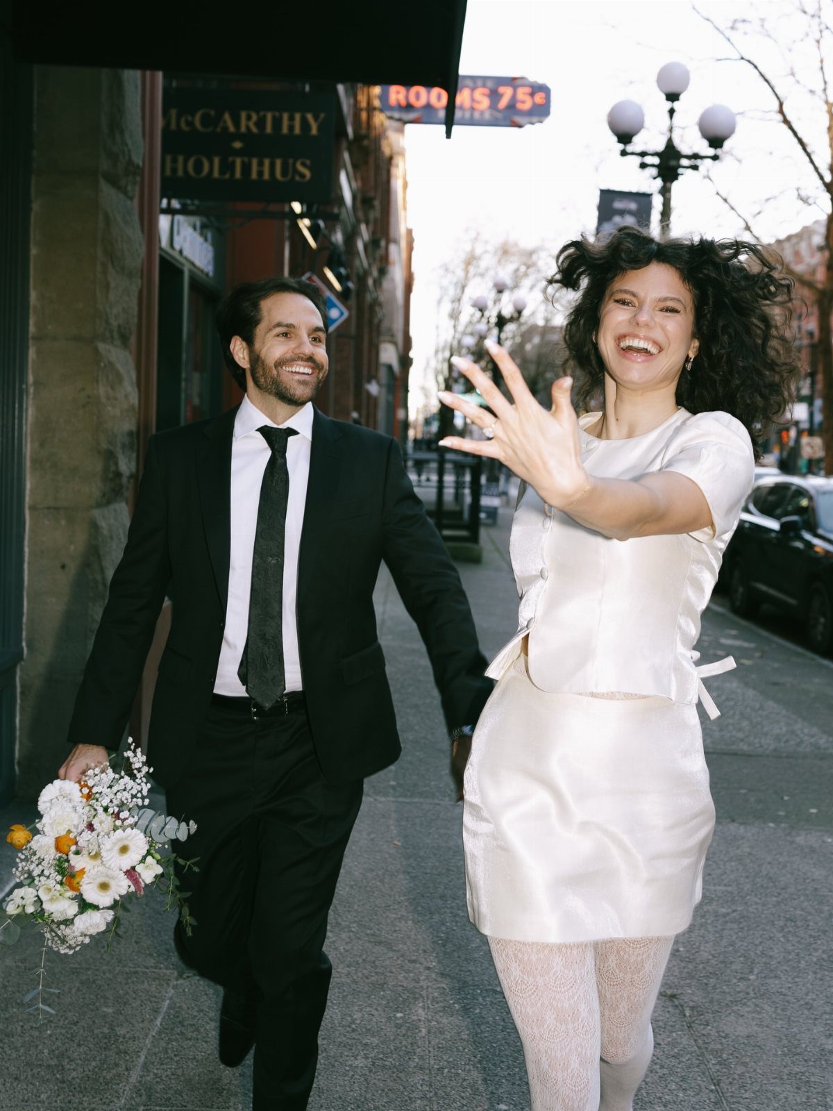 Smiling couple in wedding attire walking hand-in-hand on a city street sidewalk