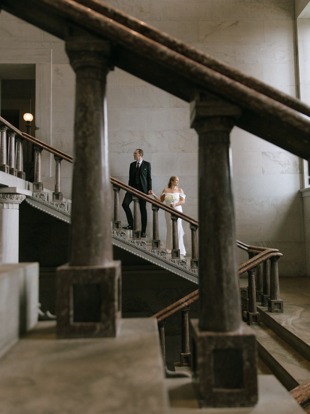 Bride and groom posing on a grand marble staircase in a classical building.