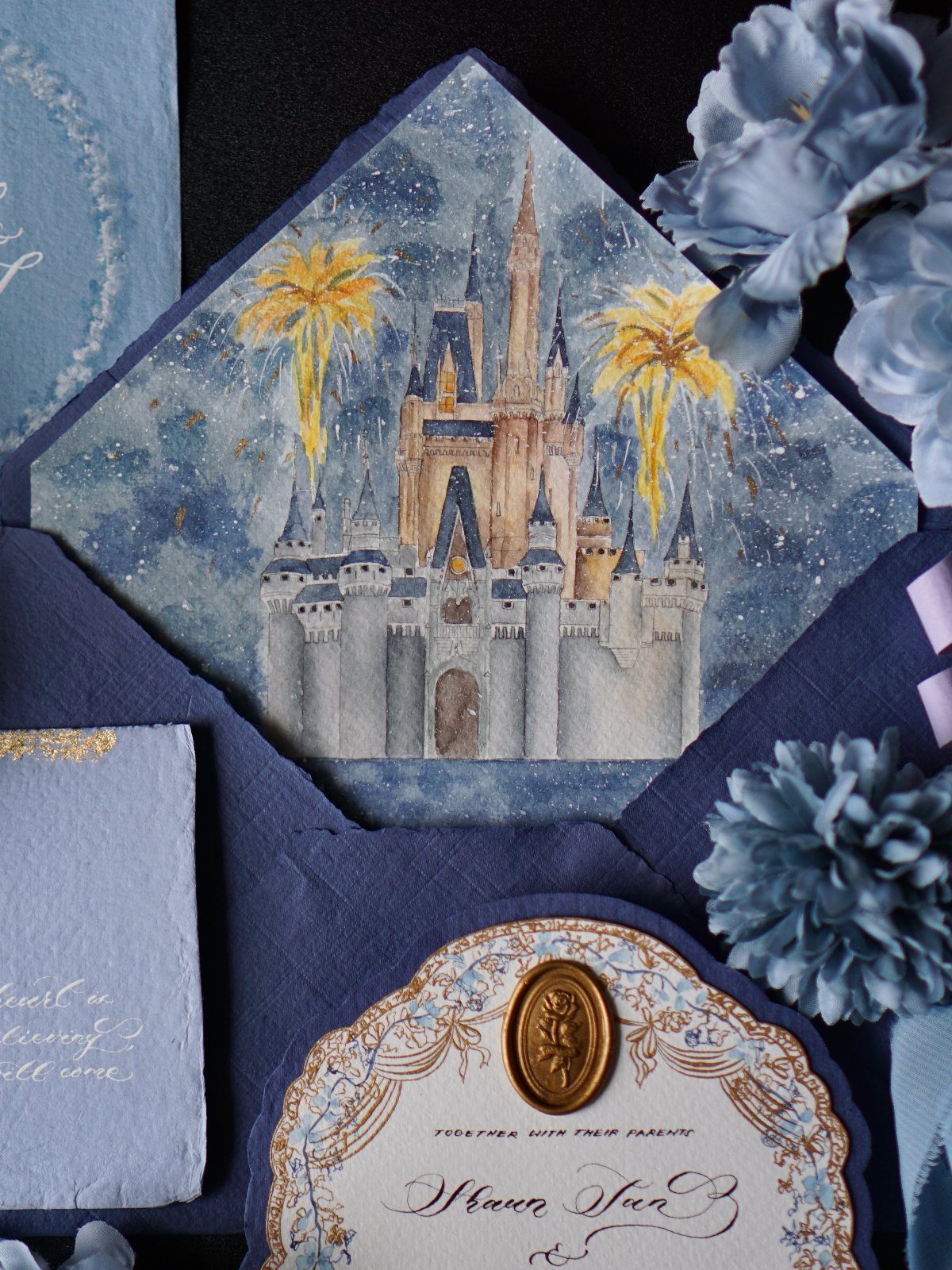 Watercolor castle wedding invitation with blue accents, wax seal, and blue flowers.