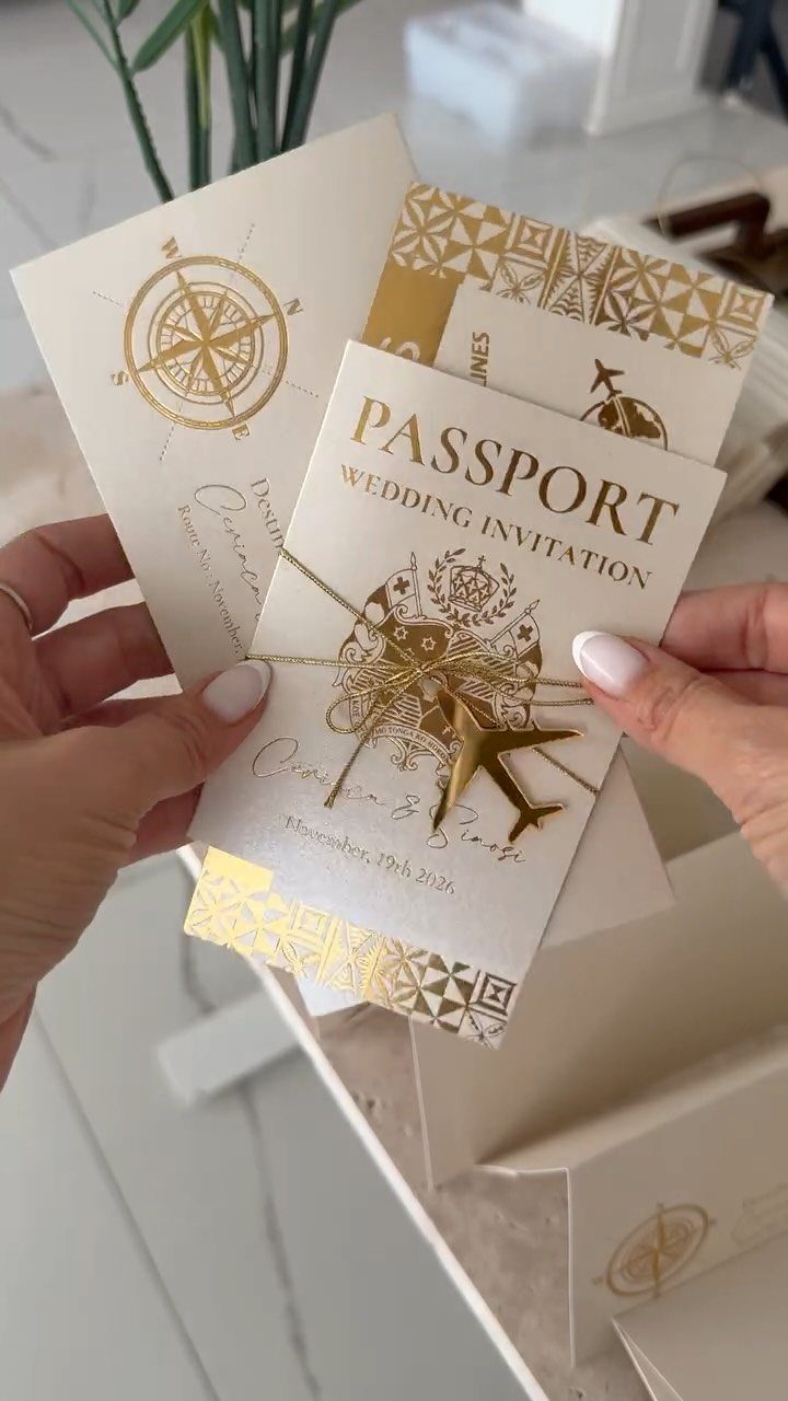 Passport-style wedding invitations with gold foil accents and a compass design held by hands.