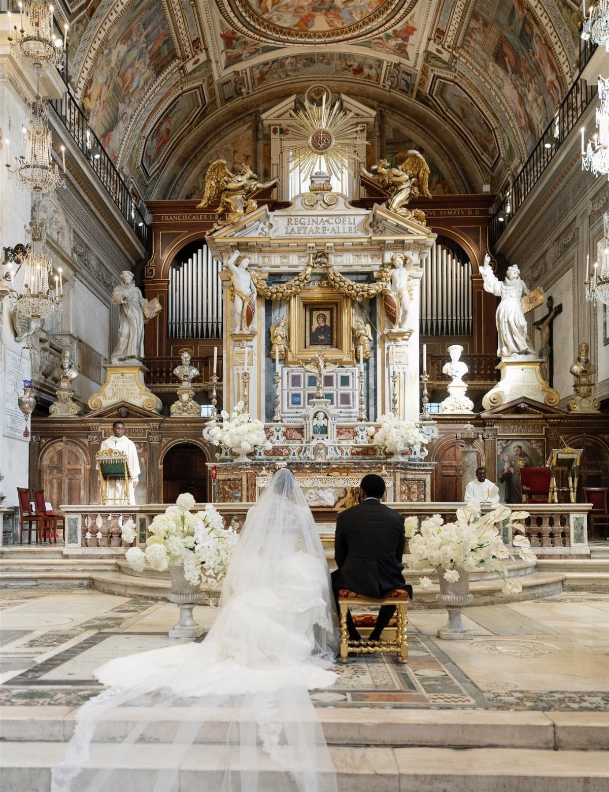 Bride and groom sitting before a lavish gold altar in a grand cathedral.
