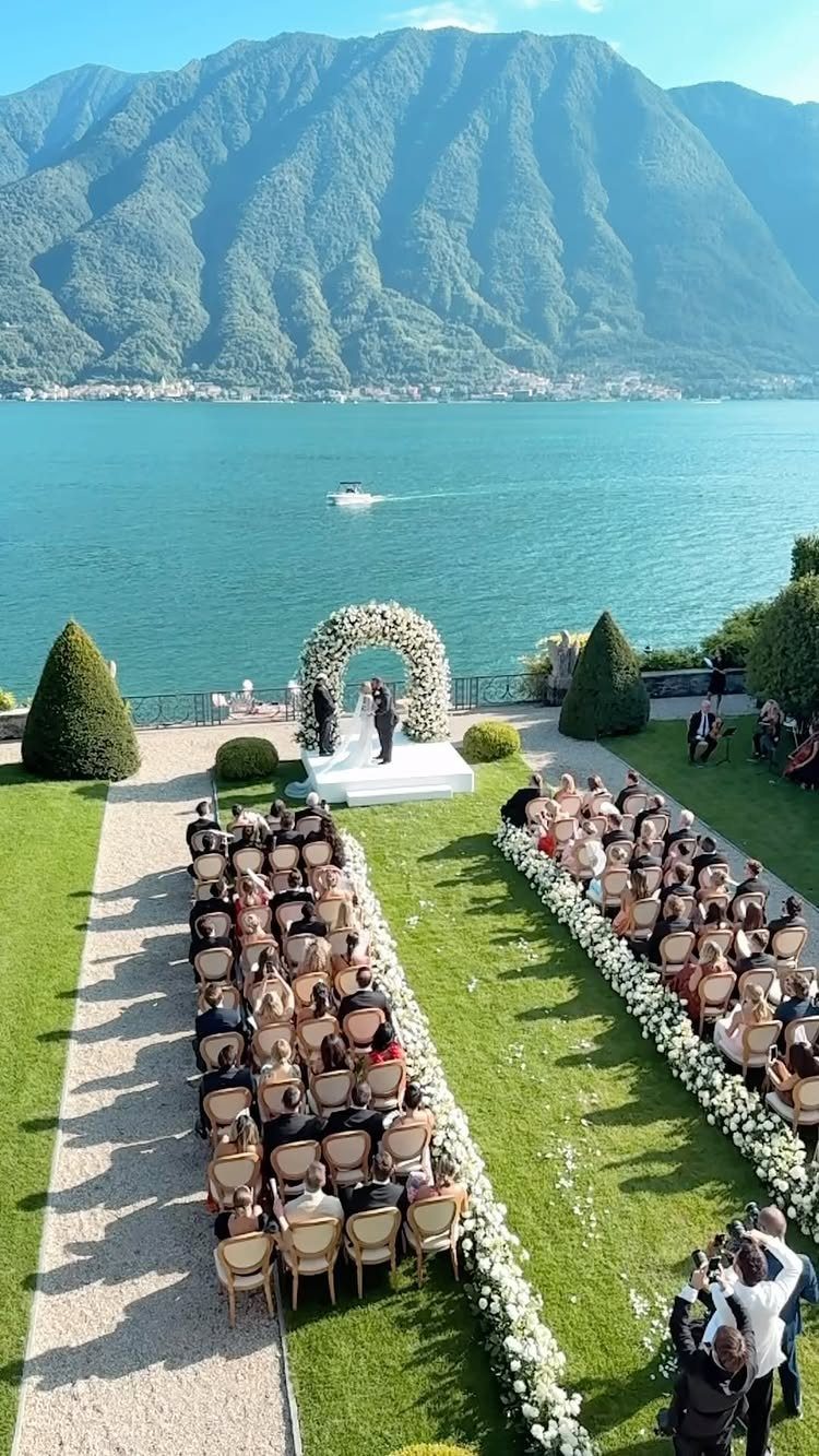 Wedding ceremony on a lakeside lawn with a white floral arch and mountains.