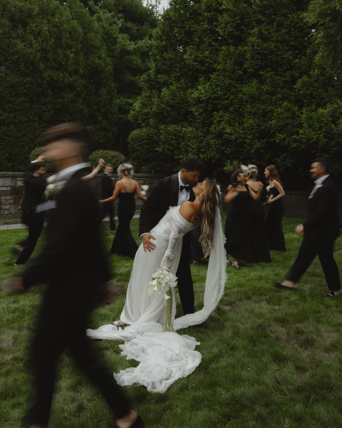 Bride and groom kissing in a garden with blurred guests in black attire.