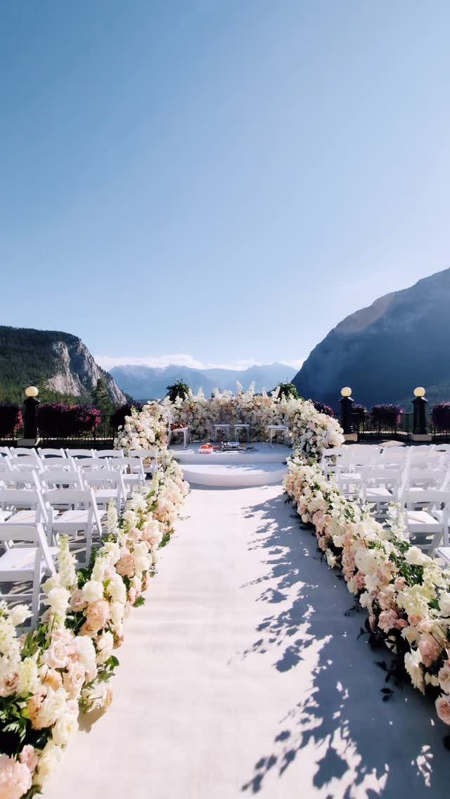 Outdoor wedding ceremony setup with white flowers and chairs against a mountain backdrop.