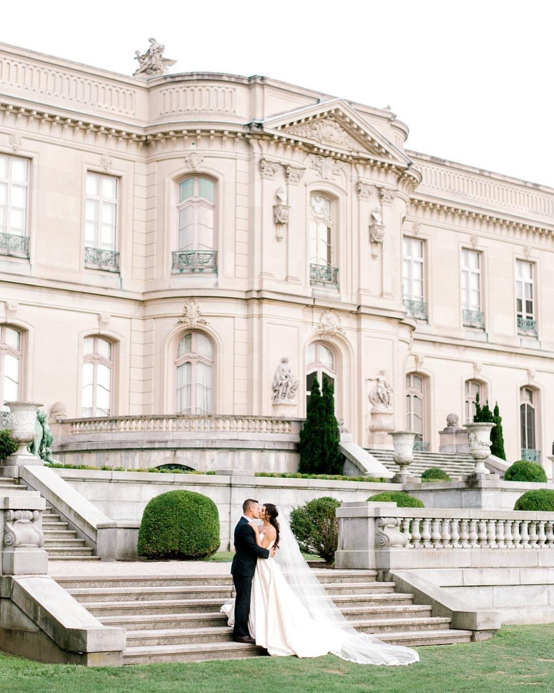 Bride and groom kissing on the stone steps of a grand neoclassical mansion.