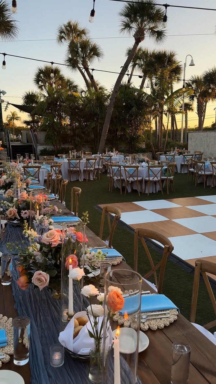 Outdoor wedding reception with decorated tables, a dance floor, and palm trees at dusk.