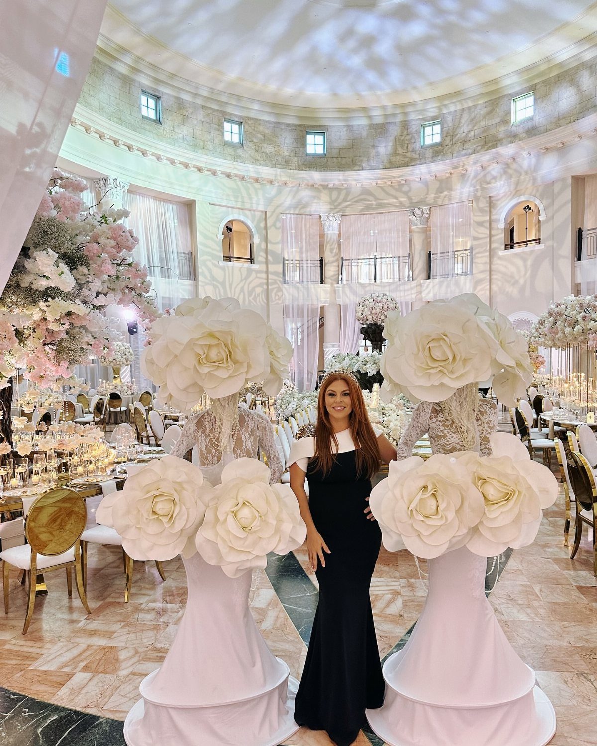 Woman in black dress posing among giant white floral sculptures in a ballroom.
