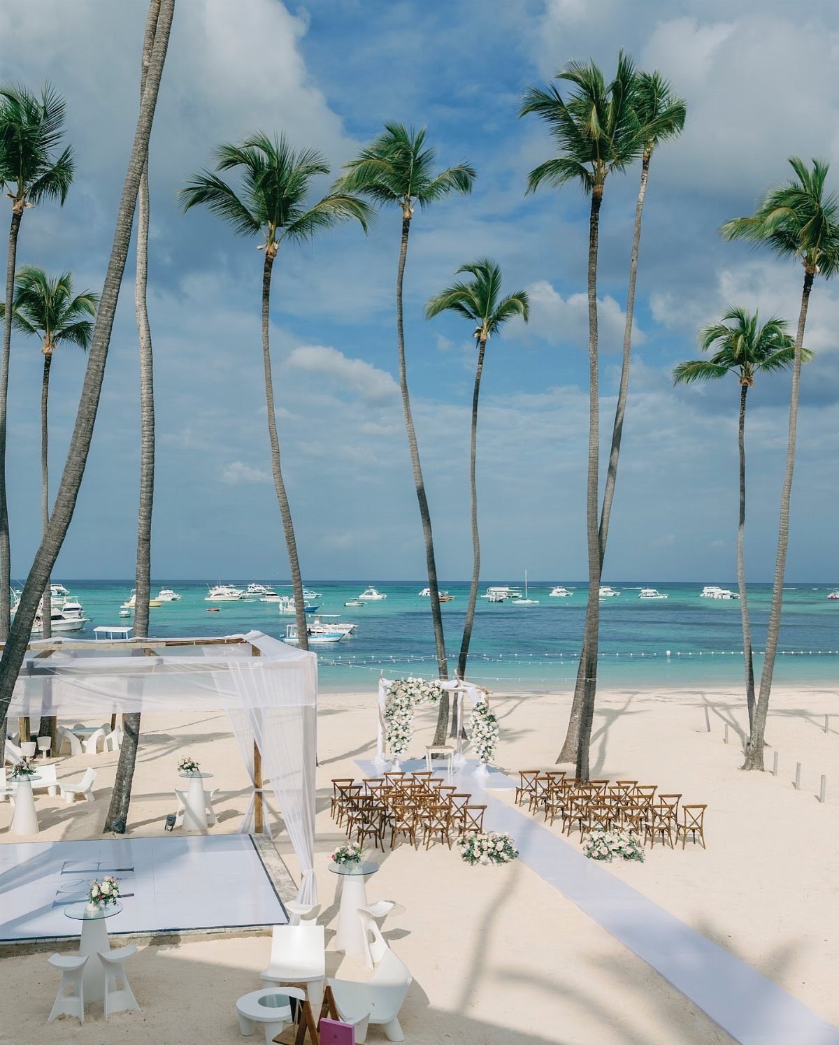 Beach wedding ceremony setup with white aisle, wooden chairs, and palm trees.