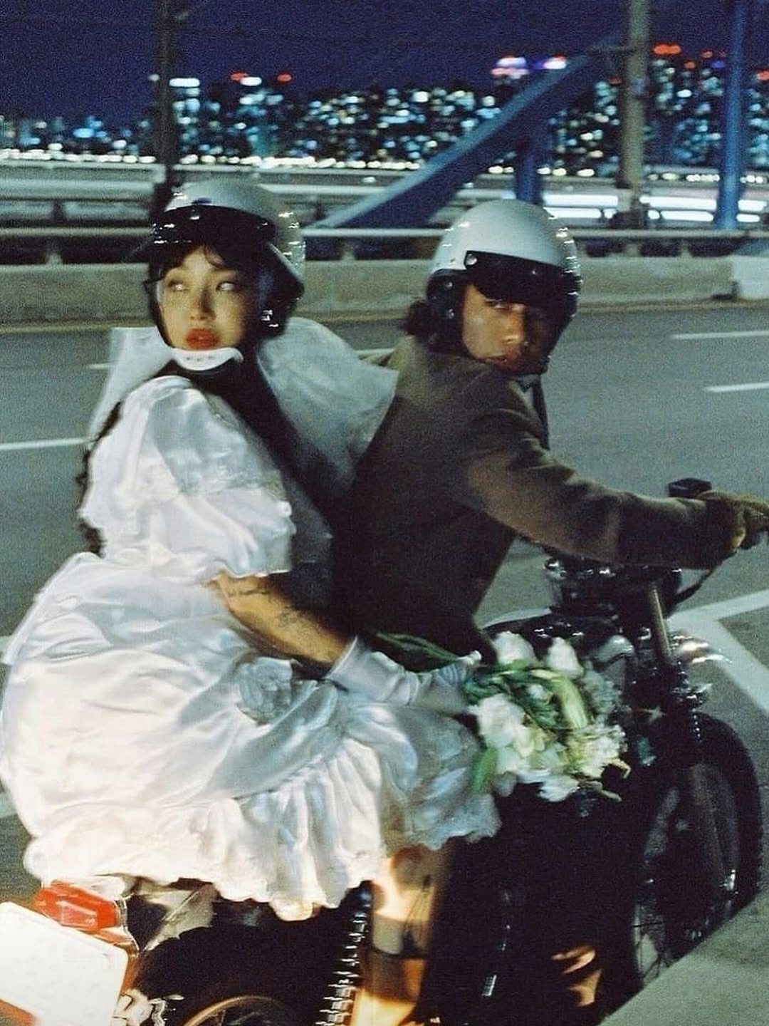 Couple in wedding attire and helmets riding a motorcycle at night near city lights