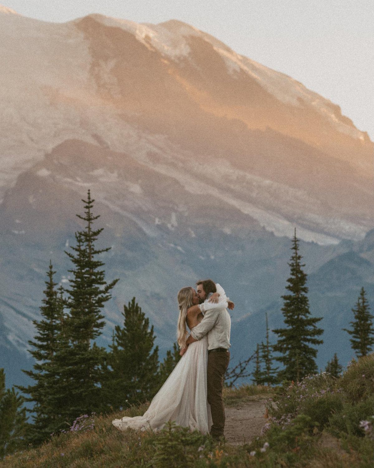 Couple embracing on a mountain ridge with a massive snow-capped peak behind them.