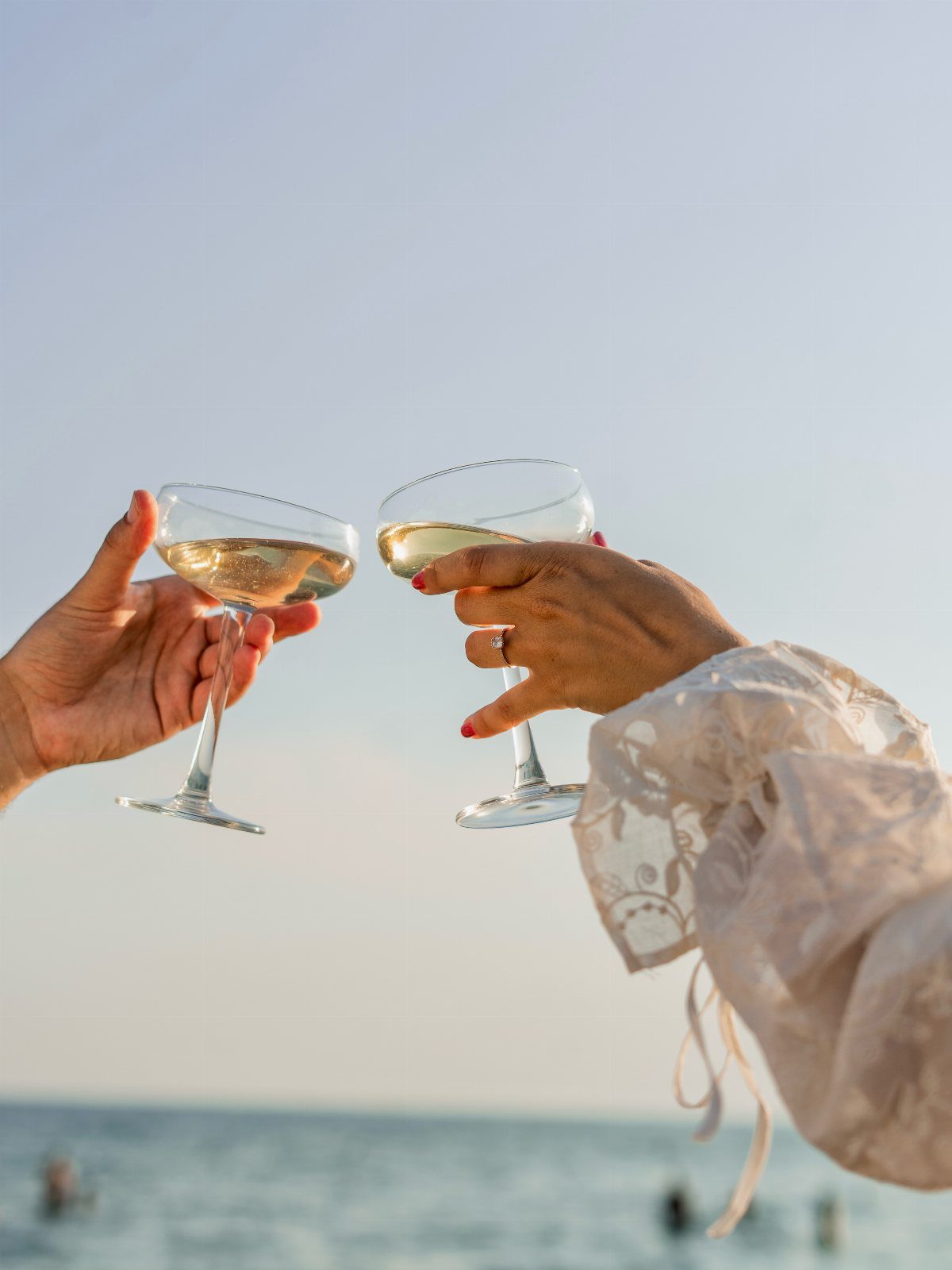 Two champagne coupes clinking in a toast against a beach and sky background.