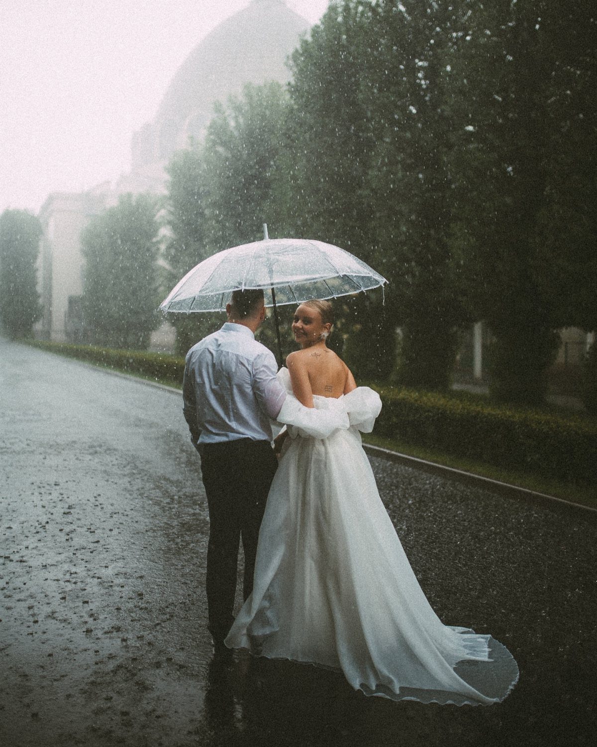 Wedding couple standing under a clear umbrella on a rainy road with trees.