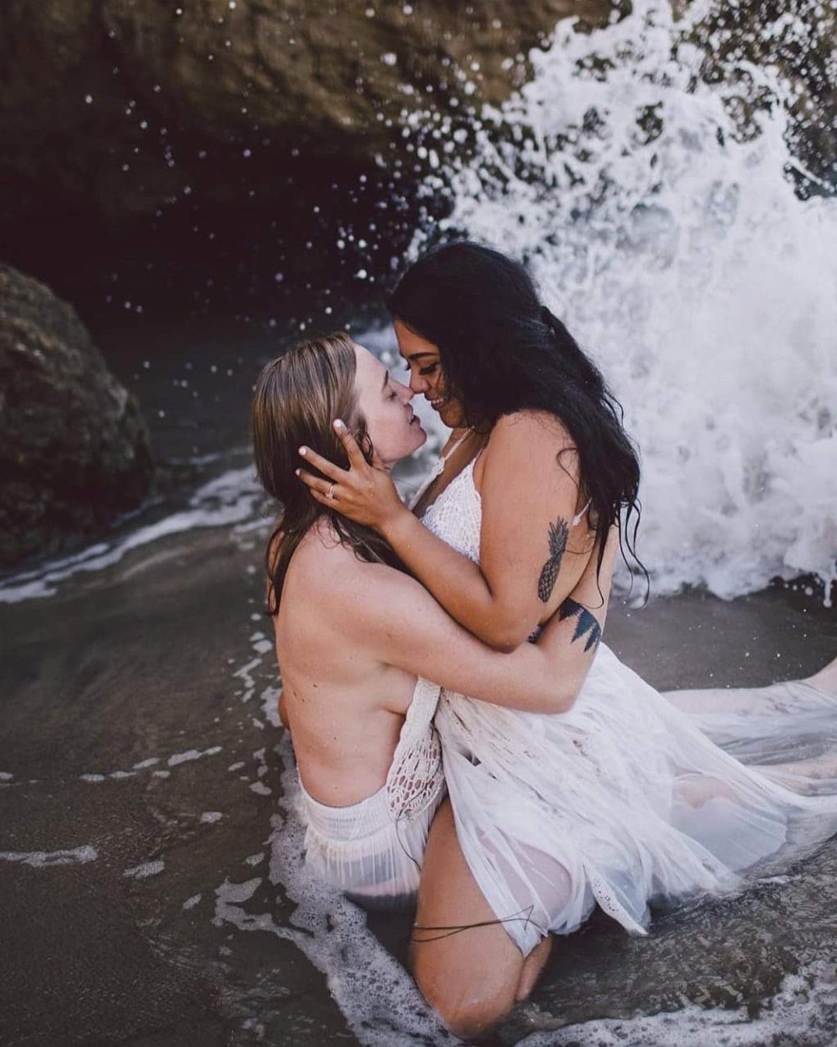 Two women in white dresses embracing in ocean surf with crashing waves.