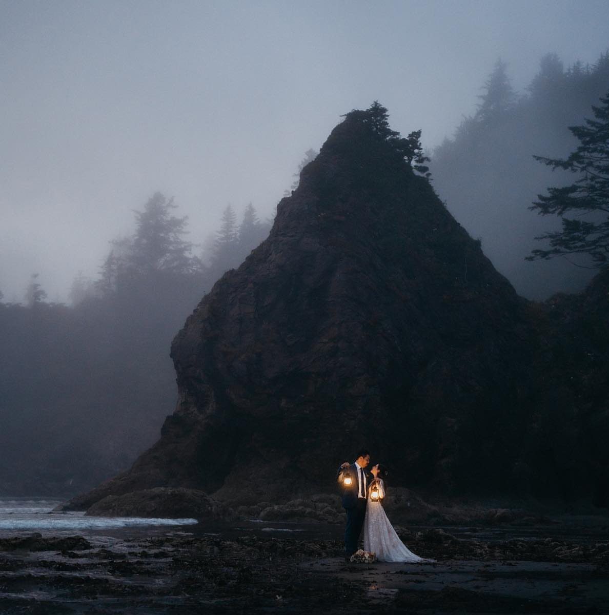 Couple in wedding attire holding glowing lanterns on a misty, rocky beach.