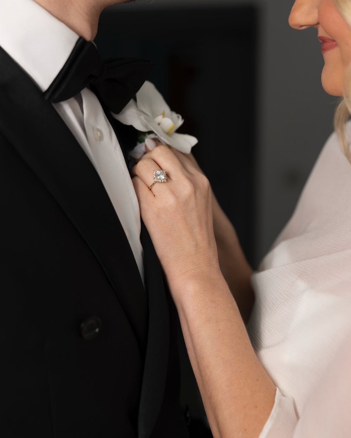 Woman's hand with diamond ring adjusting a white flower boutonniere on a tuxedo.