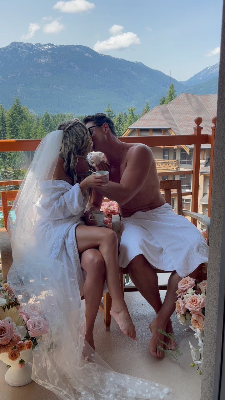 Couple in bridal attire and towels kissing on a balcony with mountain views