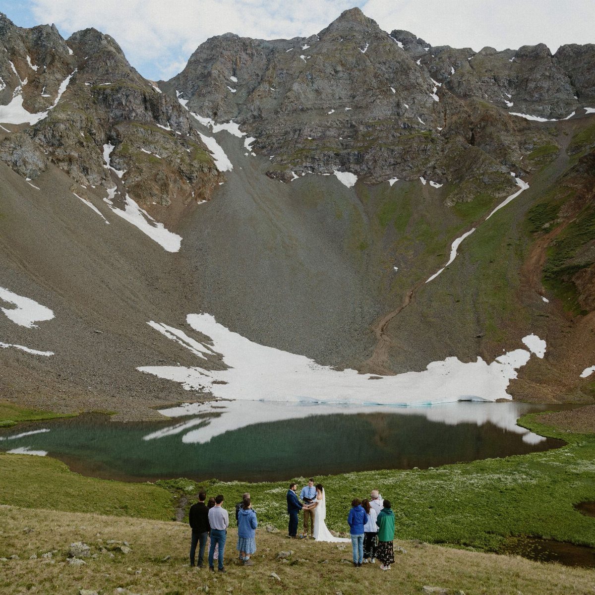 Wedding ceremony on a grassy mountain meadow overlooking a turquoise alpine lake.