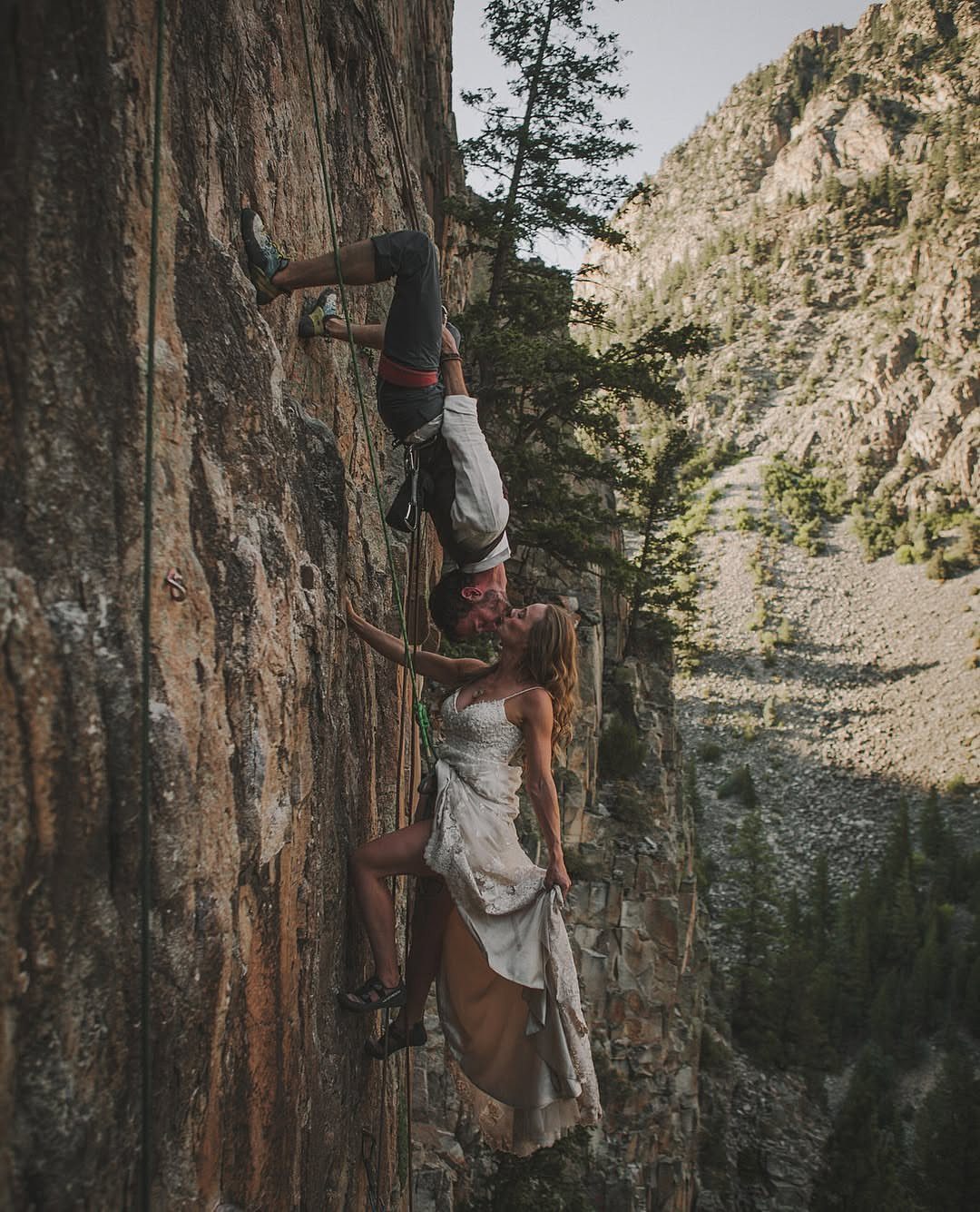 Couple in wedding attire kissing while rock climbing on a steep mountain cliff