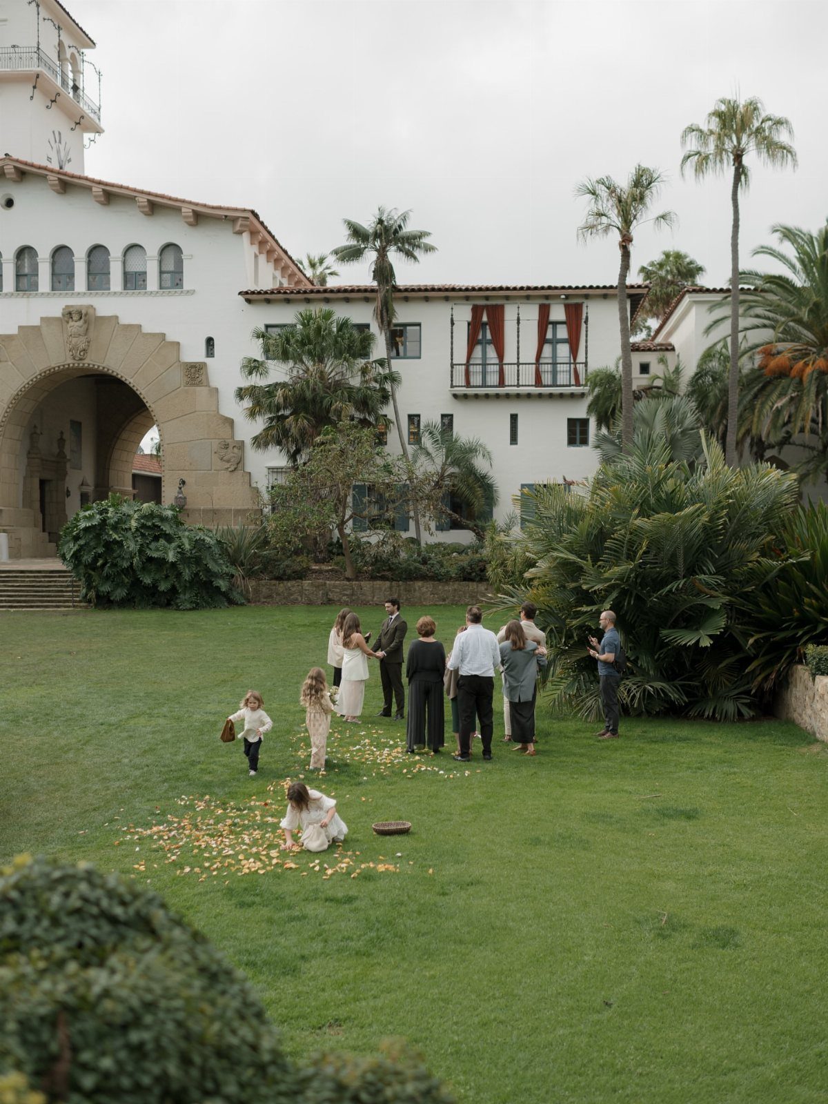 Wedding couple kissing on a green lawn surrounded by guests and flower petals.