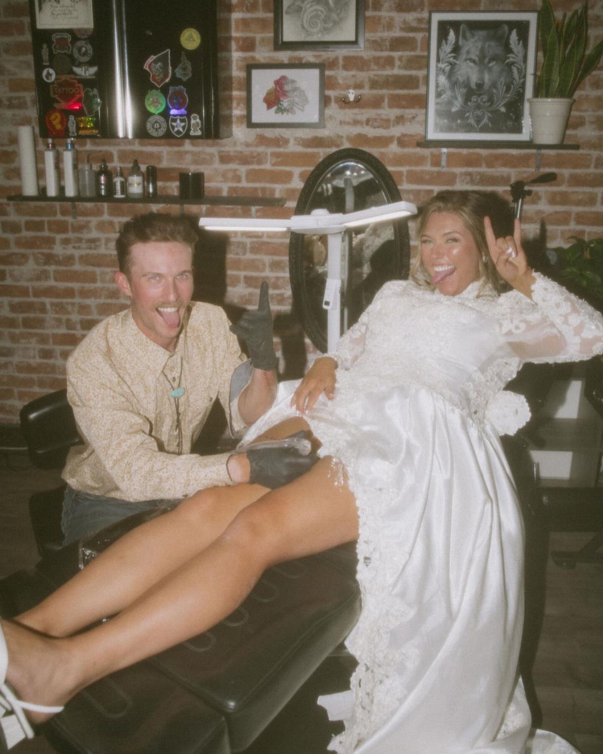 Couple in wedding attire posing playfully inside a tattoo parlor with brick walls.