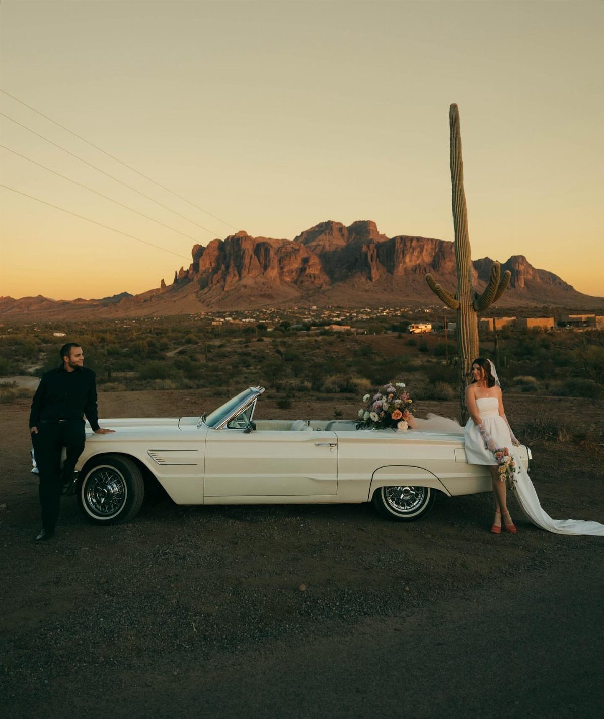 Couple in wedding attire posing with a white vintage convertible in the desert.