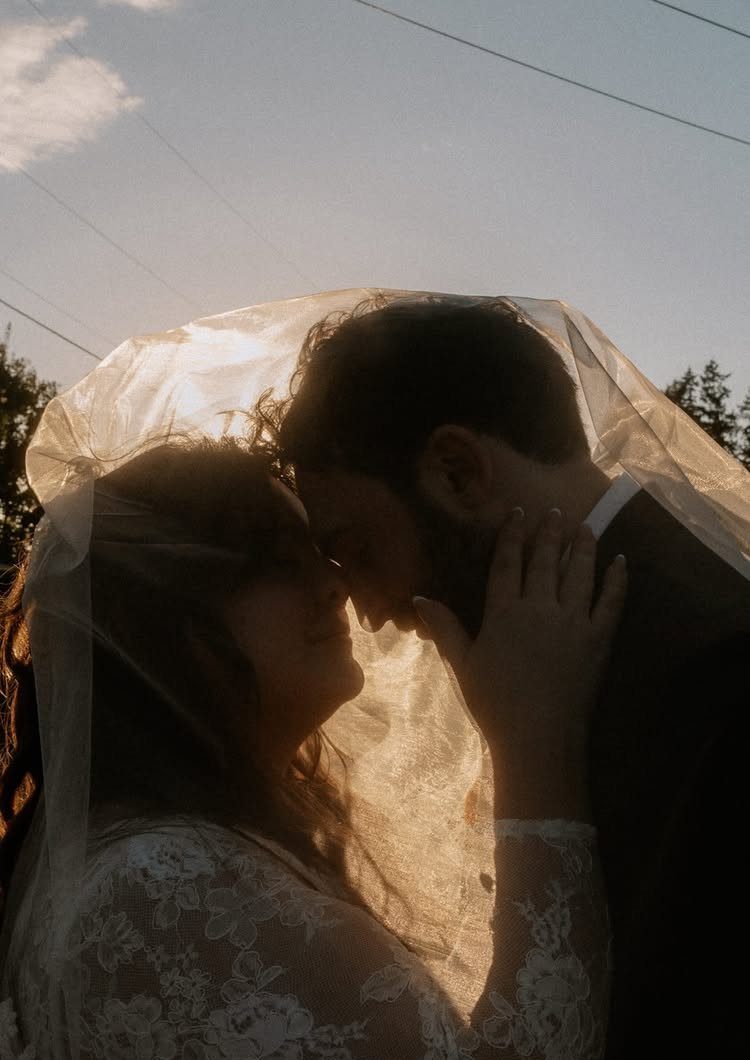 Couple touching foreheads under a white wedding veil in warm sunlight