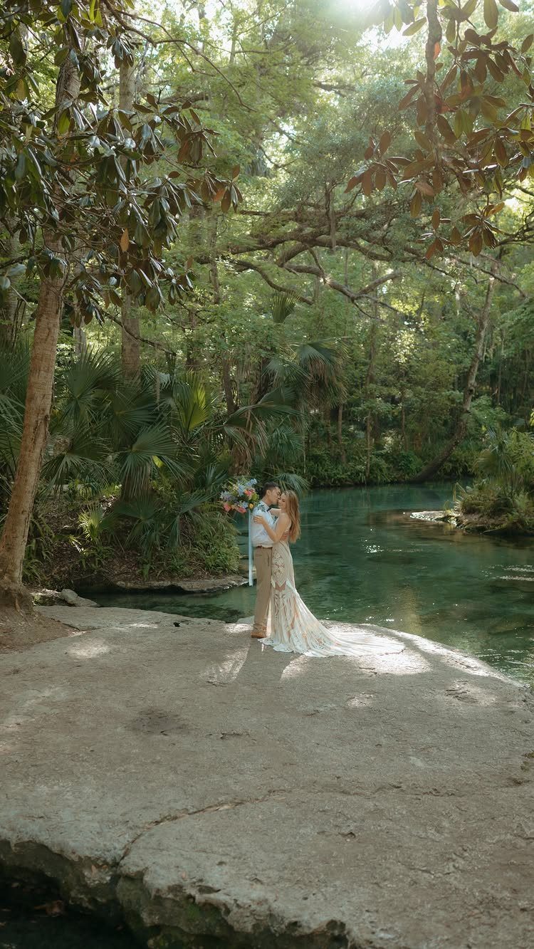 Couple in wedding attire kissing on a rocky riverbank in a lush forest