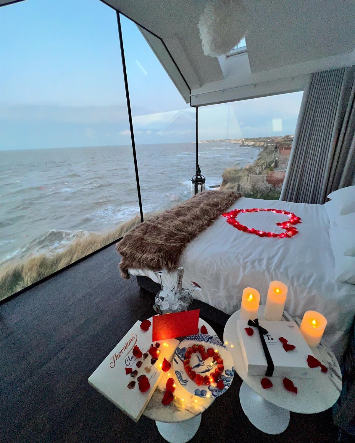 Modern glass-walled bedroom overlooking the ocean with rose petals on the bed.