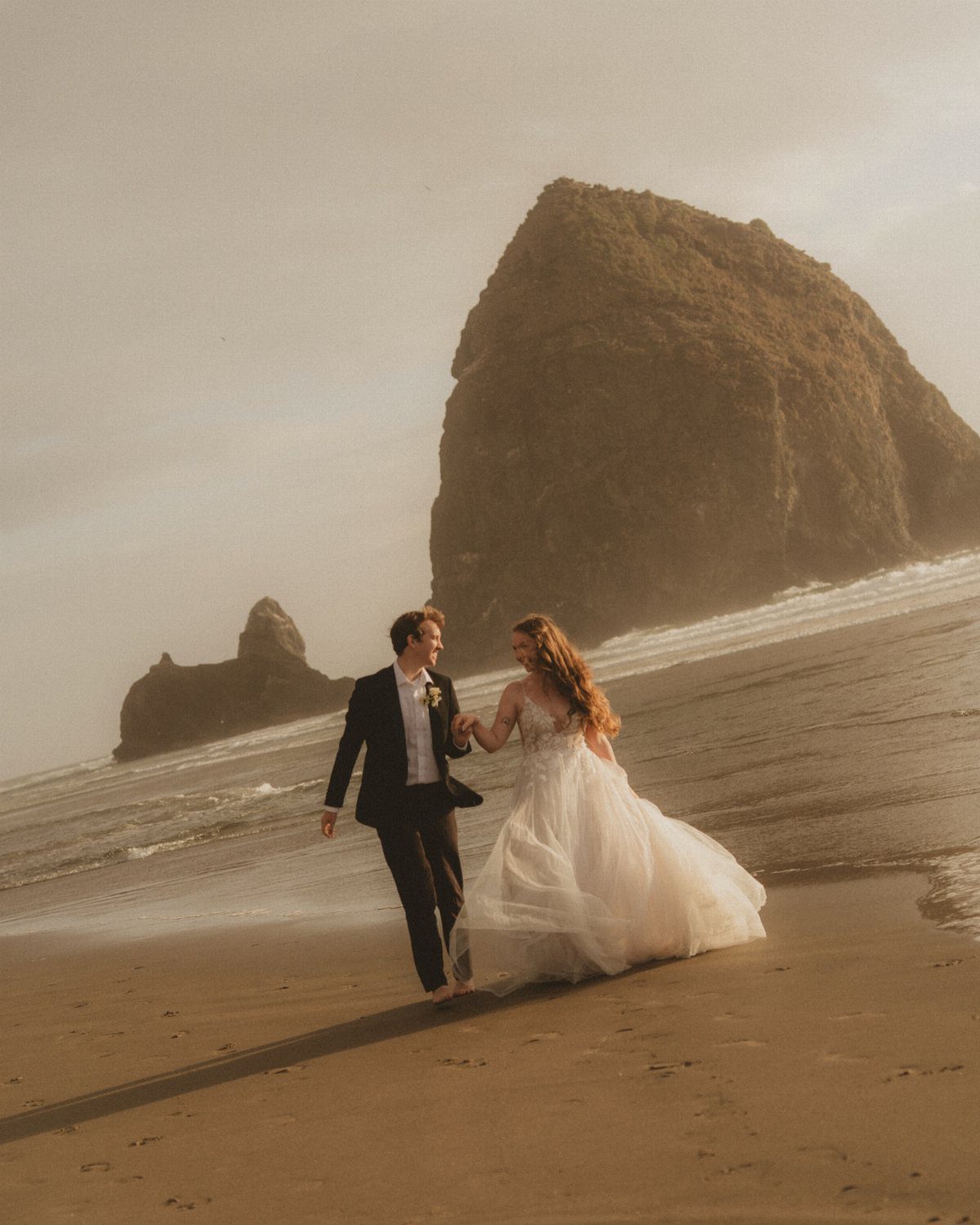 Wedding couple walking hand in hand on a beach with large rock formations.