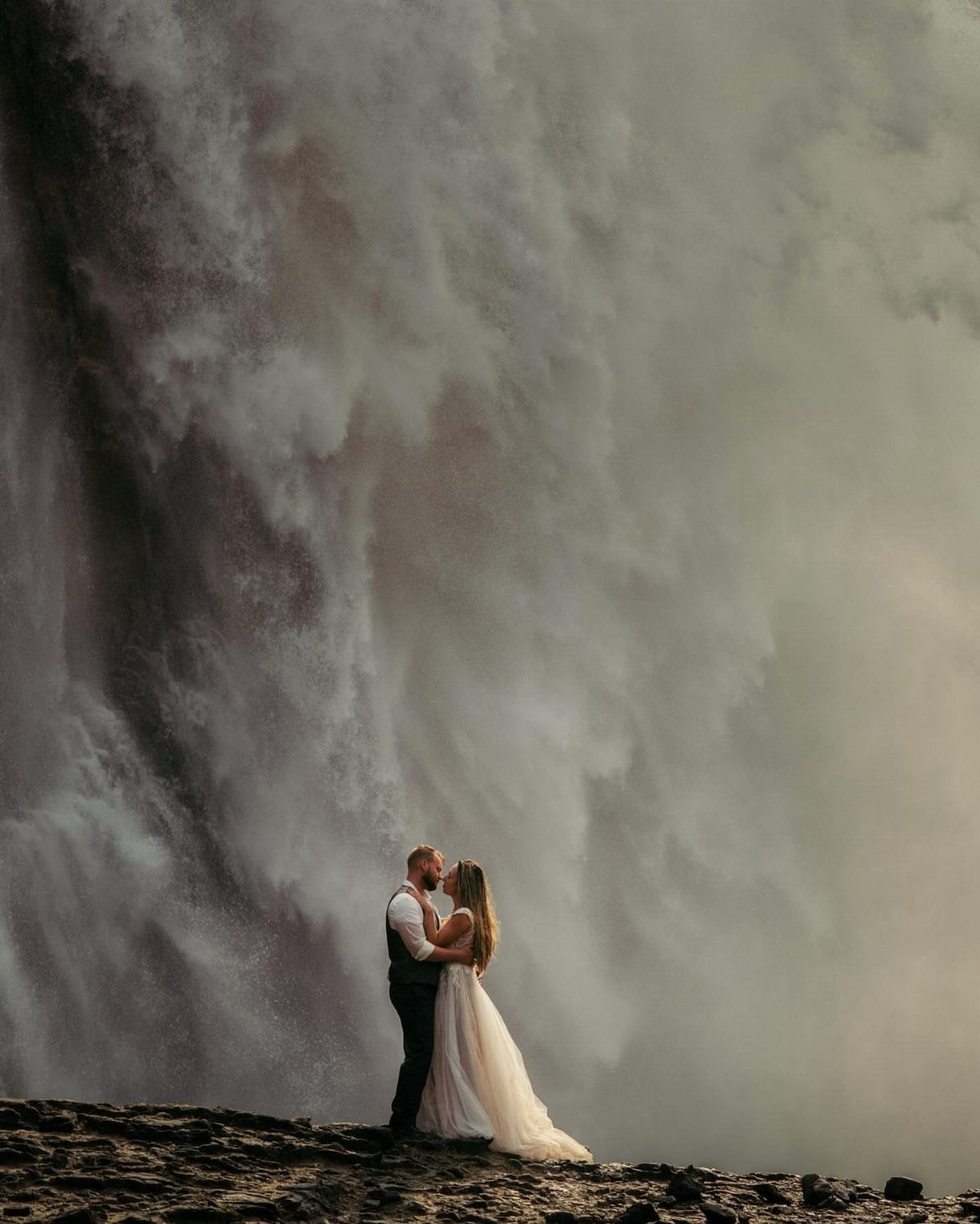 Couple in wedding attire kissing before a massive, misty waterfall on dark ground