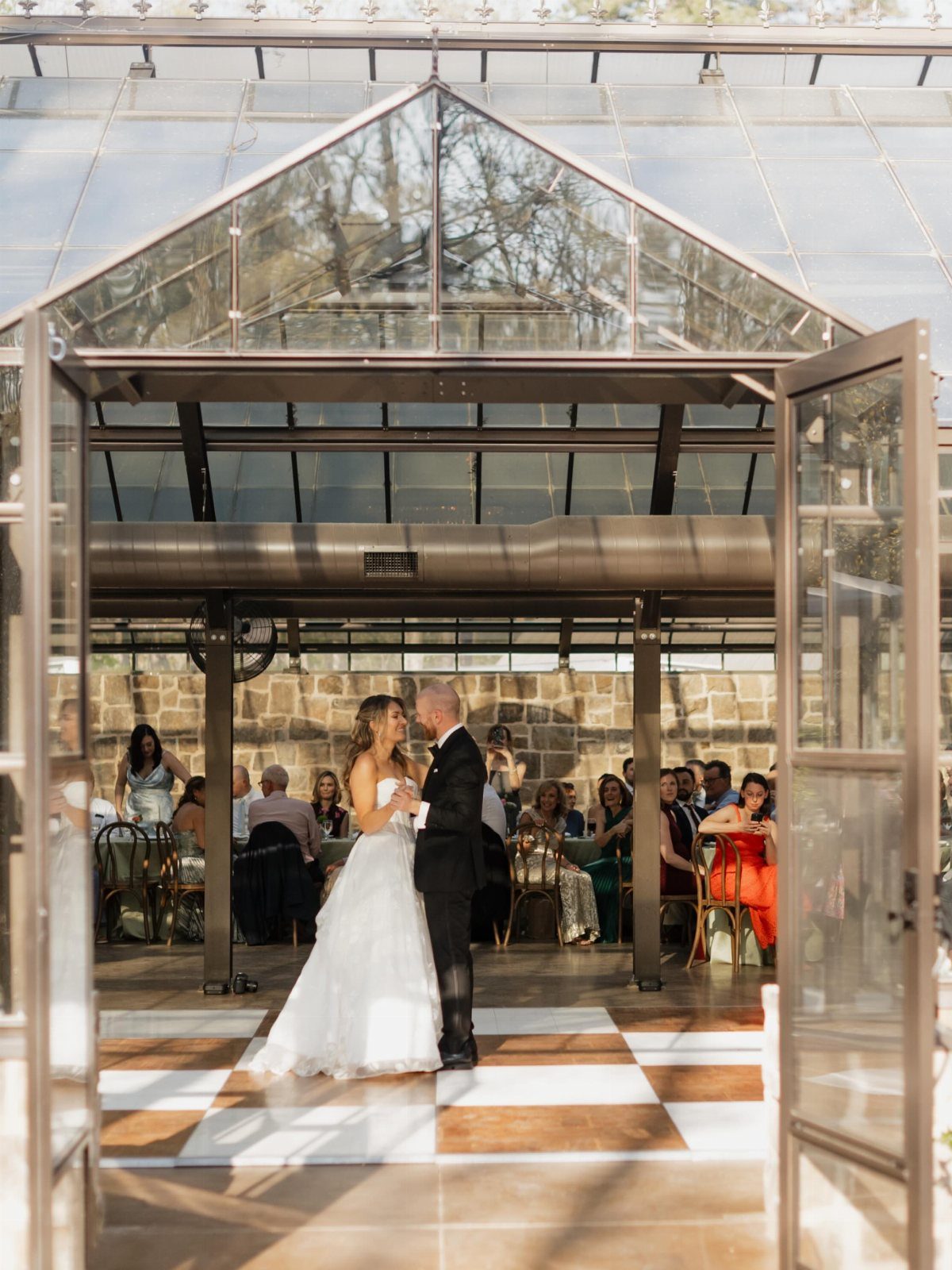 Bride and groom dancing inside a glass house venue with seated wedding guests.
