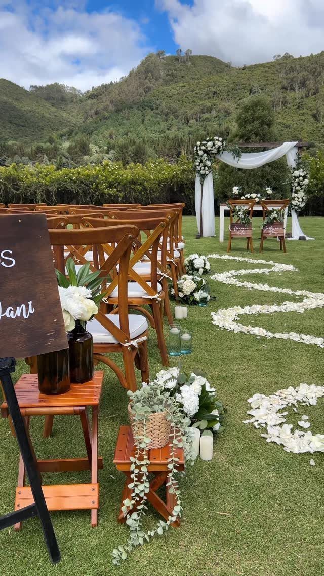 Outdoor wedding ceremony setup with wooden chairs and white floral altar against mountains.