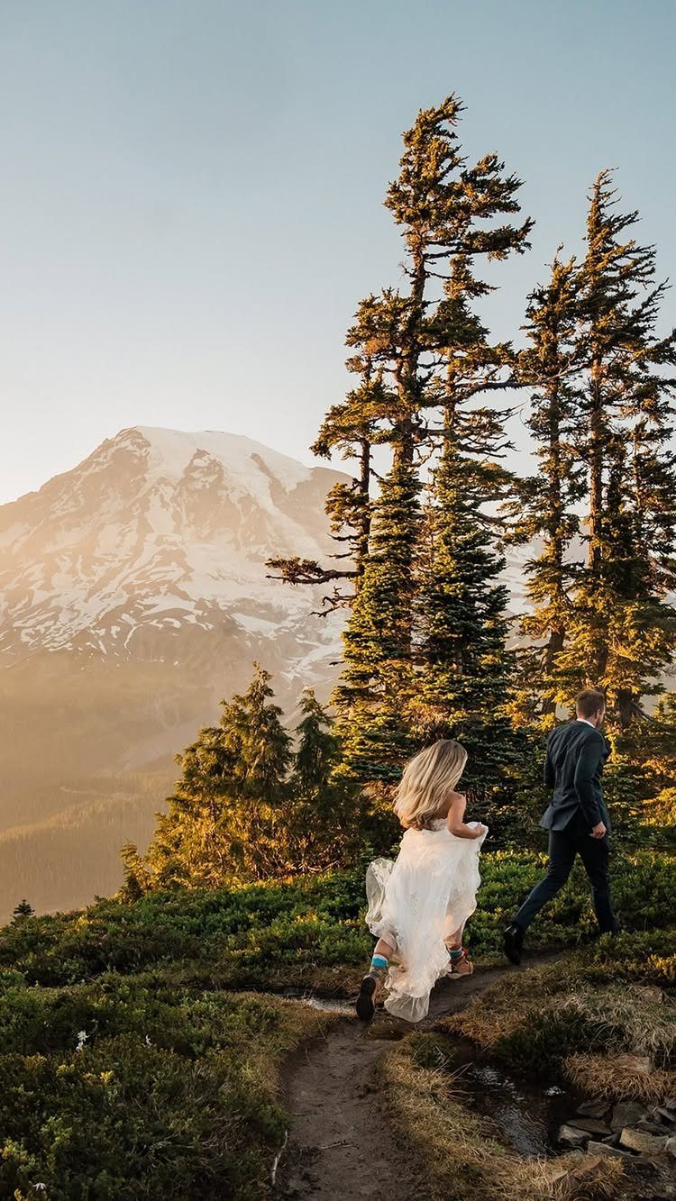 Bride and groom walking on a mountain path with snow-capped peaks behind them.