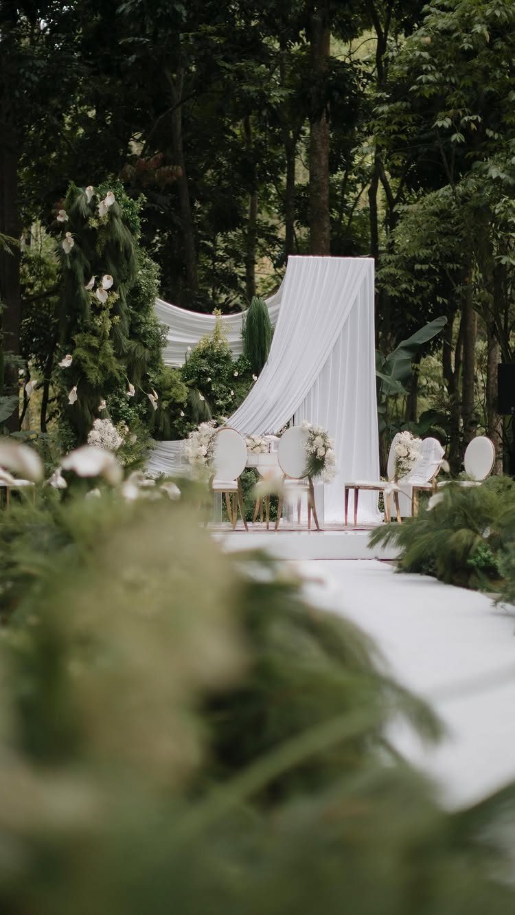 White wedding ceremony setup with fabric backdrop and floral arrangements in a forest