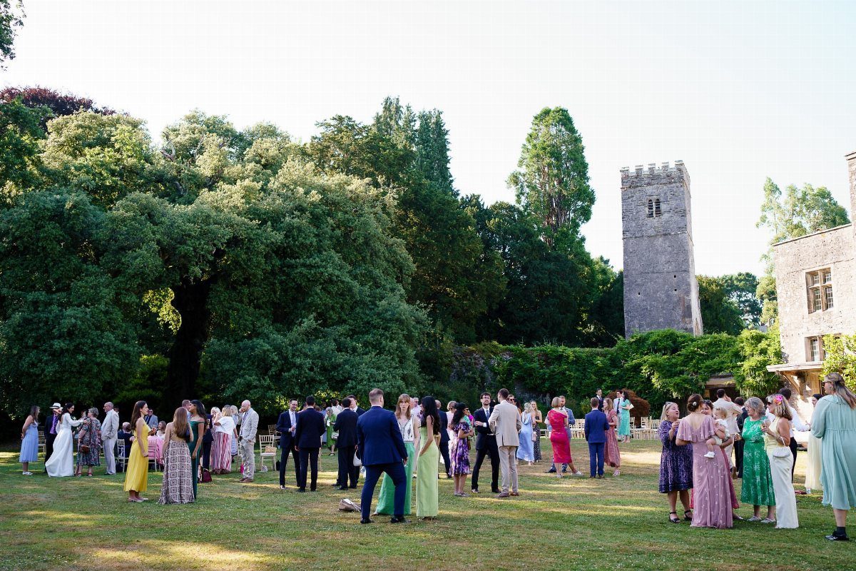 Wedding guests mingling on a grassy lawn beside a stone castle tower and trees.