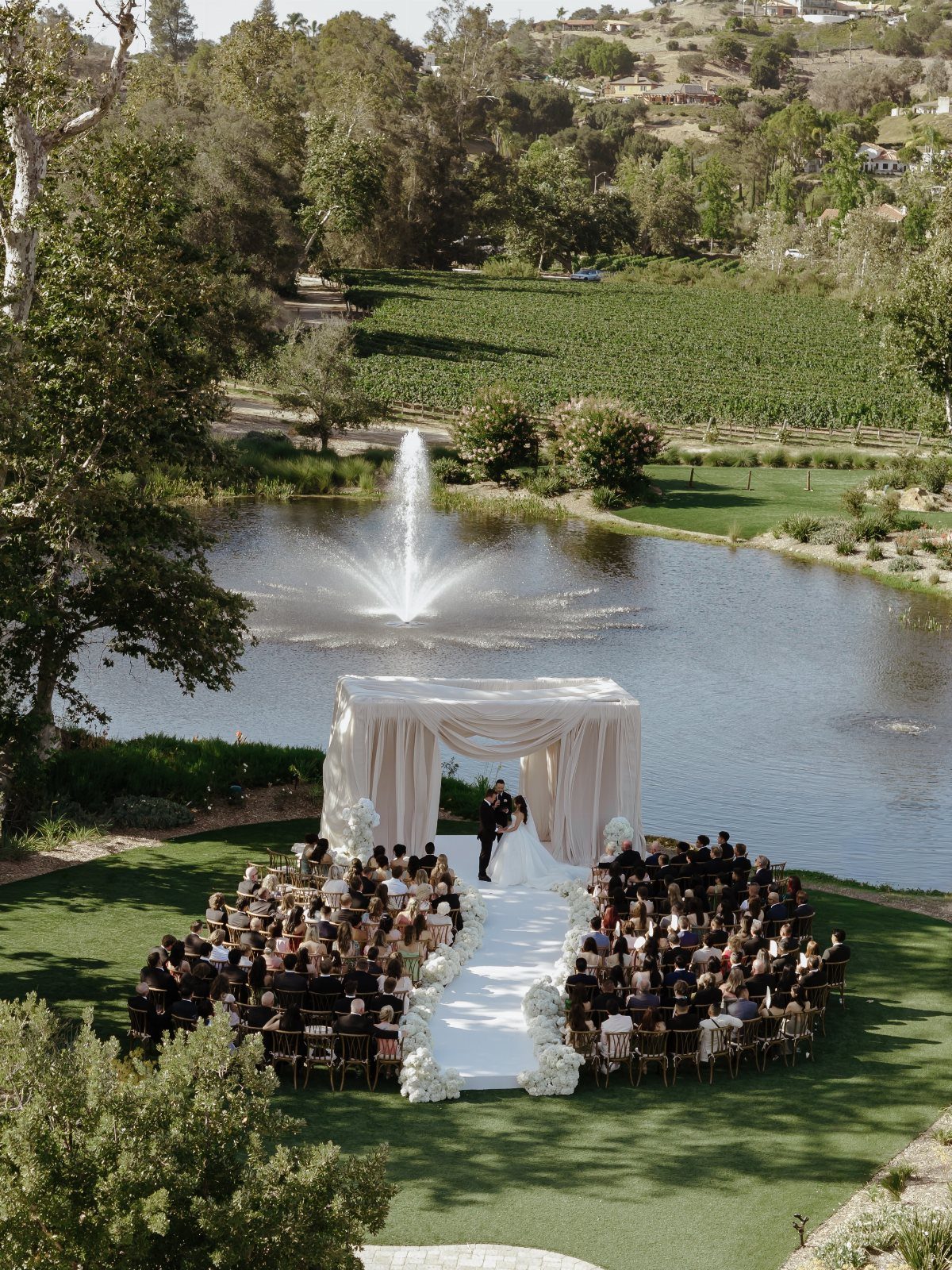 Wedding couple under a white canopy with a flower-lined aisle by a pond.