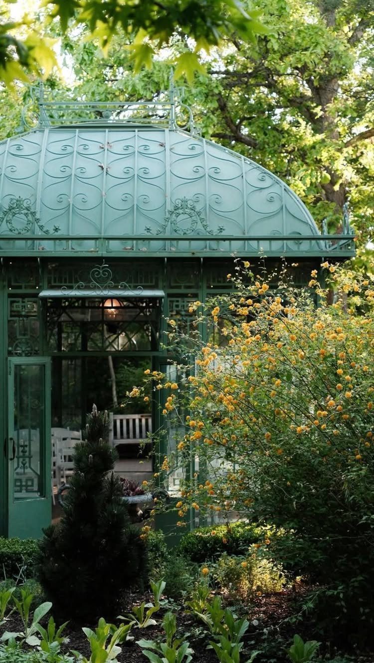 Green ornate metal conservatory dome surrounded by lush trees and yellow flowers.