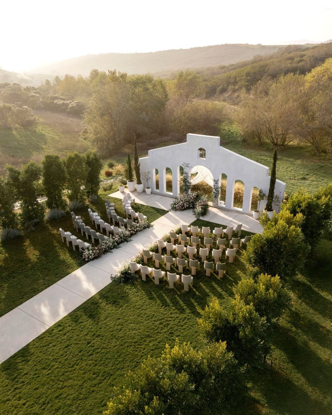 Outdoor wedding ceremony setup with white chairs and arched structure on a grassy hillside.