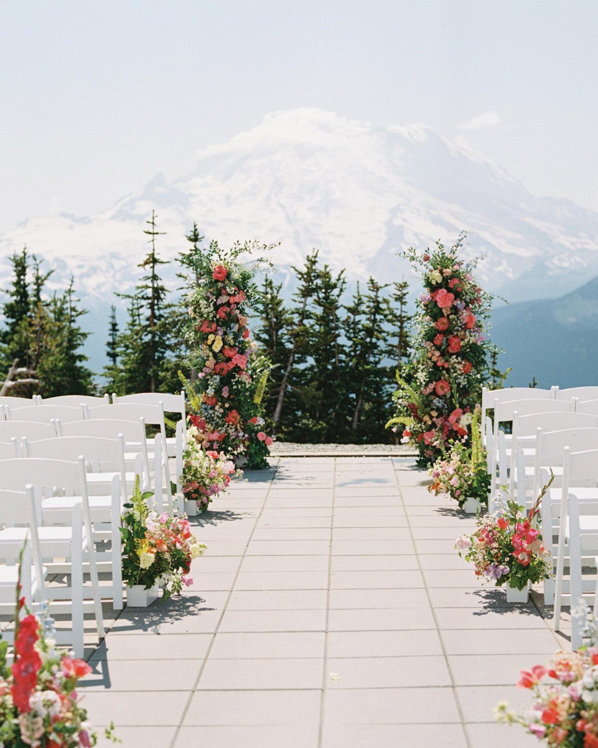 Wedding ceremony setup with white chairs and floral pillars facing a snow-capped mountain.