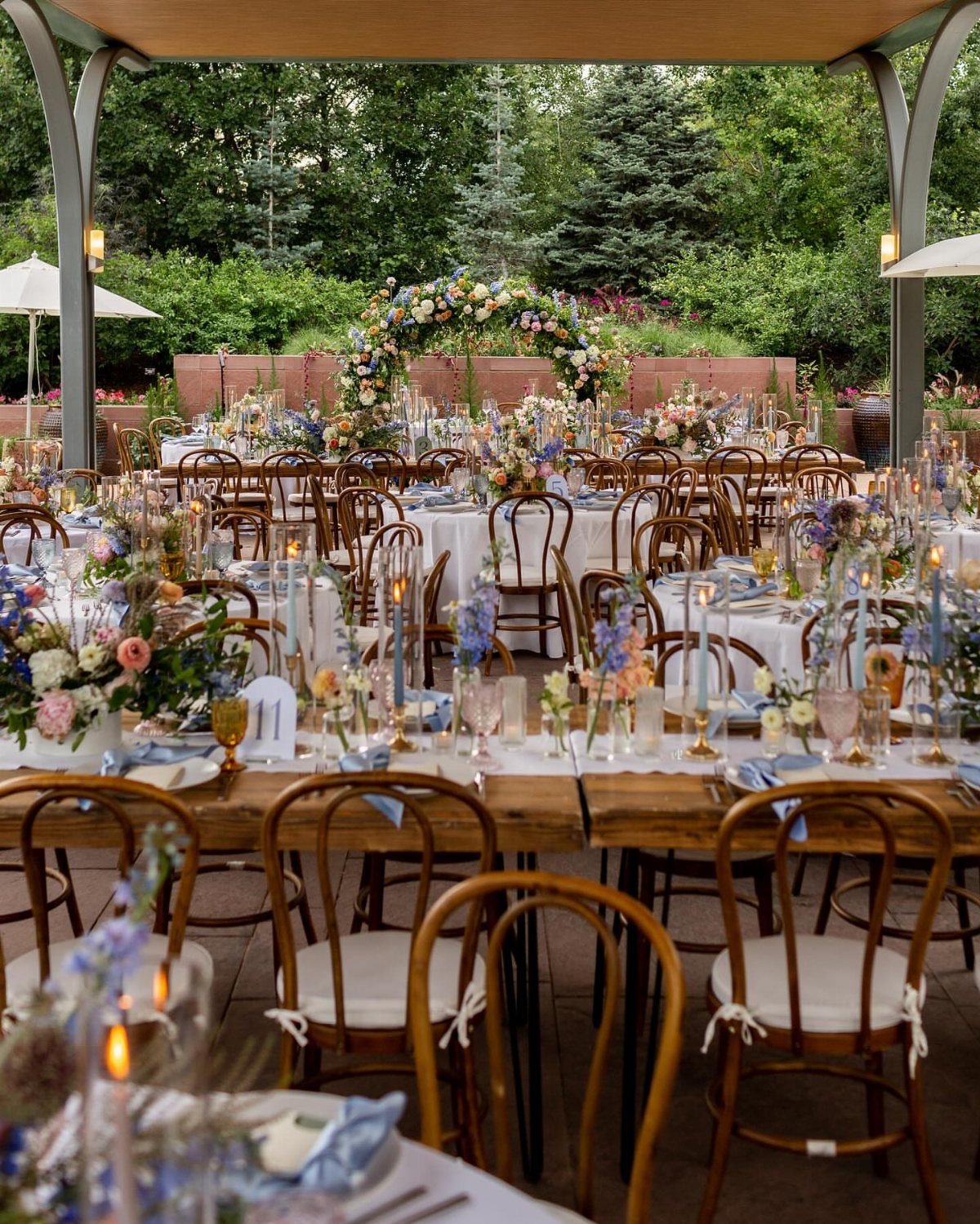 Outdoor wedding reception with wooden tables, blue floral centerpieces, and a floral arch.