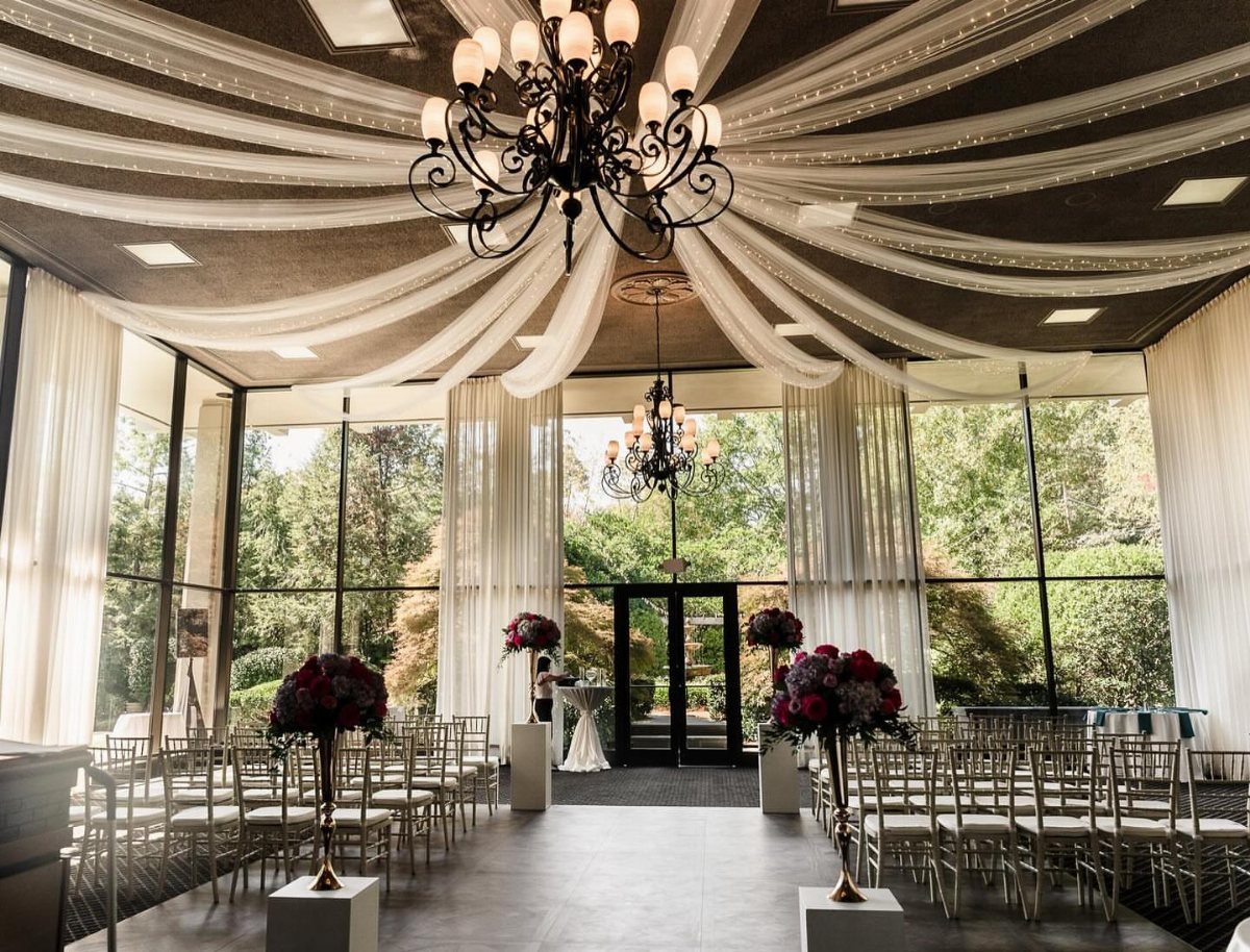 Wedding ceremony setup with white ceiling drapes, chandeliers, and silver chairs facing glass walls.