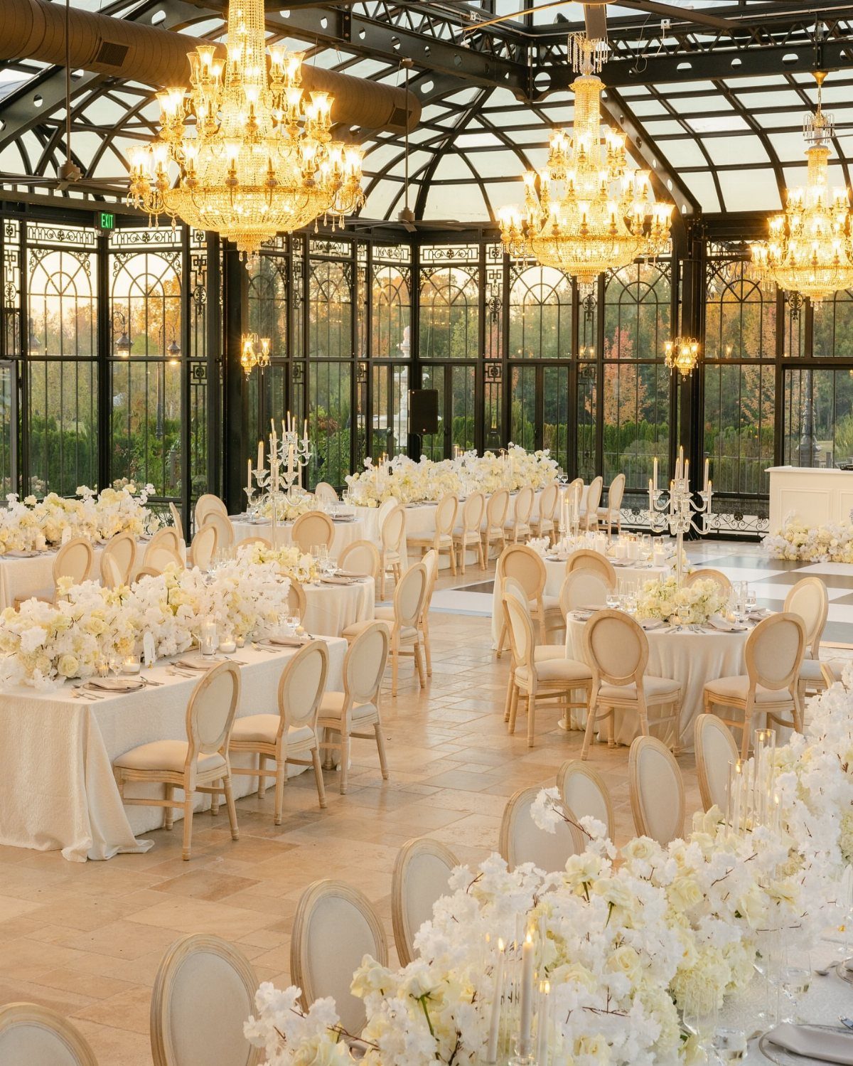 Elegant glass-walled event space with crystal chandeliers and white floral wedding table settings.
